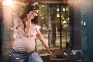 Young smiling pregnant woman typing on laptop while communicating over phone
