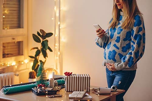 Young woman checking her phone in the middle of gift wrapping for the Holidays when pregnant