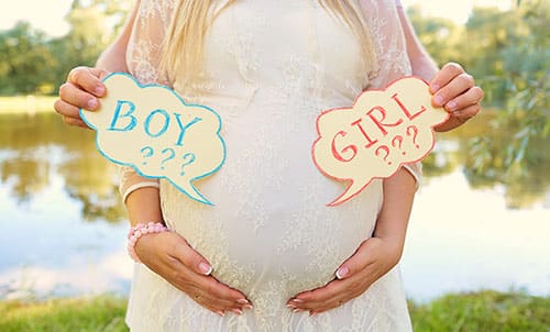 Pregnant woman cradles her belly while her husband holds signs reading 'boy???' and 'girl???'