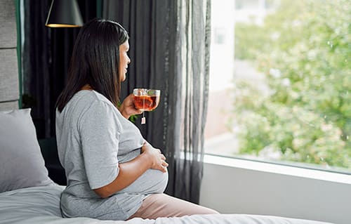 Pregnant woman seated on her bed looking outside while drinking tea
